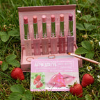 A pink display box holding six MISS ROYAL lip balm tubes in assorted pink shades, with a pink front card showing strawberry graphics and Russian text, set on artificial green grass against a white backdrop.