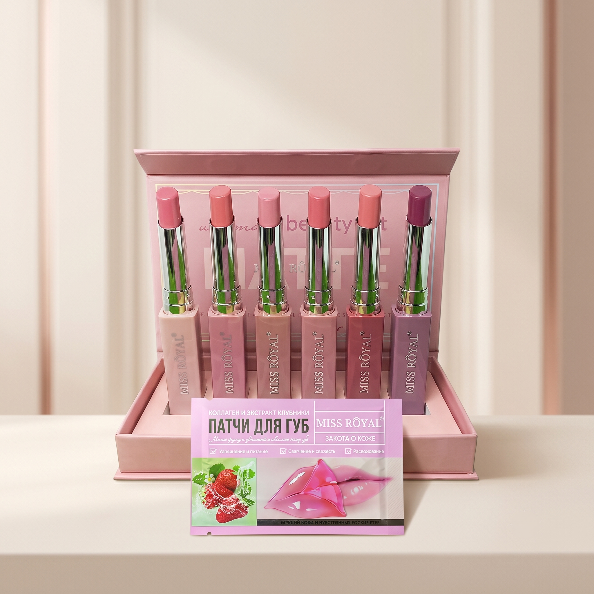 A boxed set of six MISS ROYAL lipsticks in assorted pink, peach, and mauve shades, displayed in a pink presentation box with a small pink lip treatment packet in front on a light neutral surface.