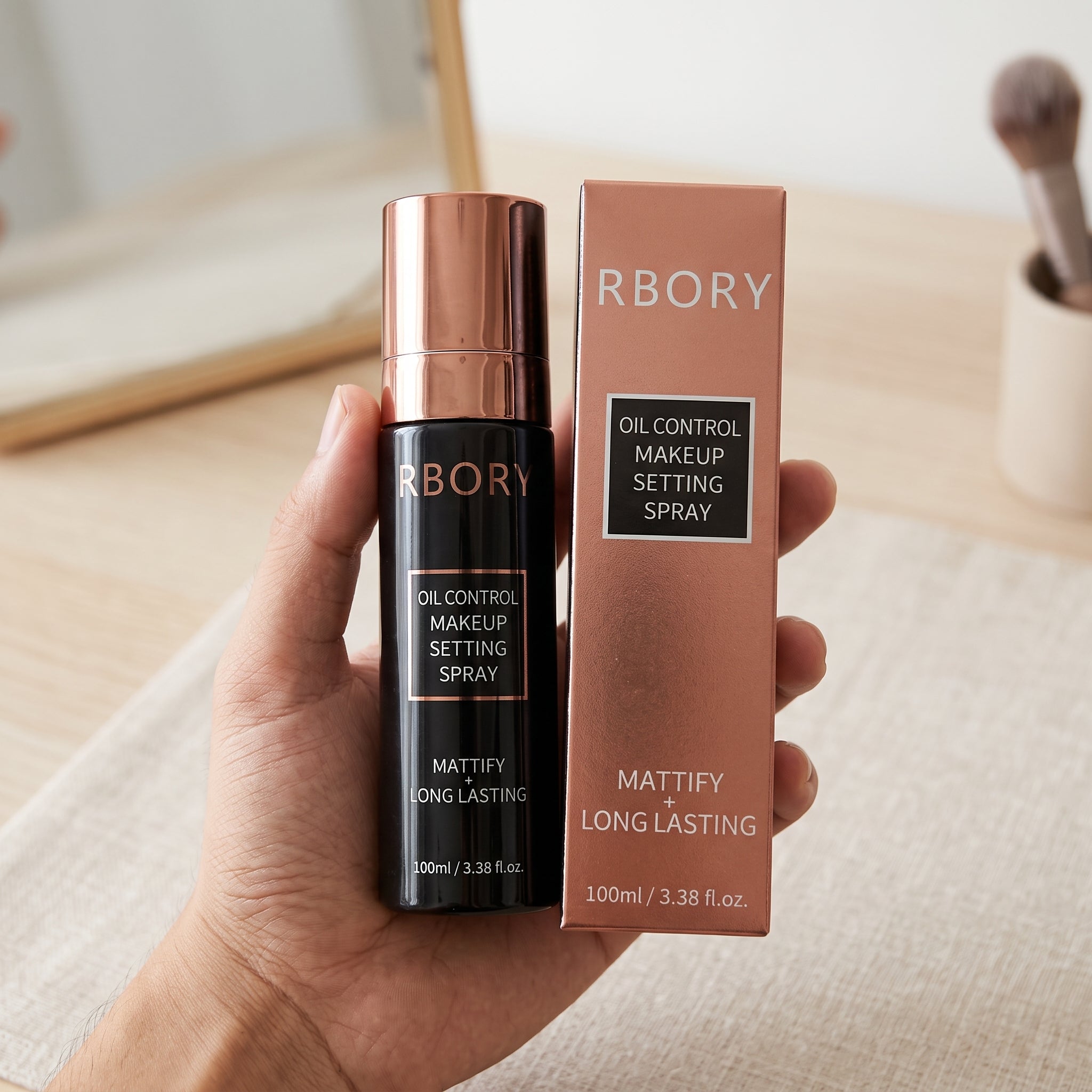 A hand holds a bottle of Burbory Decontrol Makeup Setting Spray next to its rose gold box, with shelves of other cosmetics in the background.
