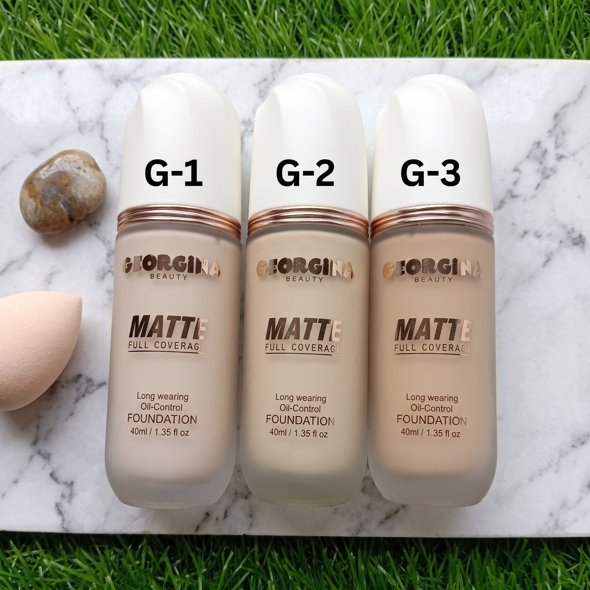 Three bottles of Georgina London Matte Full Coverage Foundation in shades G-1, G-2, and G-3, arranged side-by-side on a green grass background.