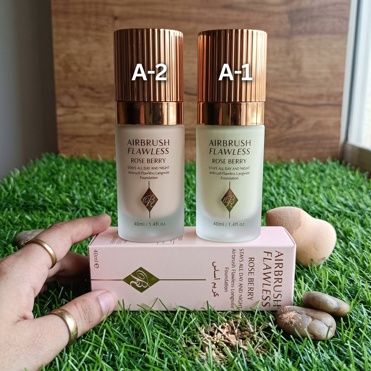 Two bottles of Charlotte Tilley Airbrush Flawless makeup in Rosé de Jóy, labeled A-2 and A-1, standing on a cream-colored product box against a light blue background with artificial grass at the bottom.