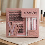 A false eyelash kit by showkiss, containing multiple rows of black false eyelashes on a pink card, a pink brow pencil, a pink mascara tube, and a pink tweezzer, arranged in a white tray on a green artificial grass background.