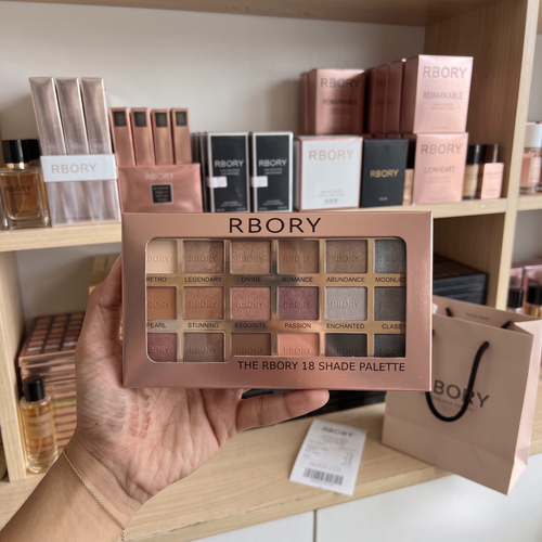 A hand holds a square makeup palette box labeled RBORY 18 SHADE PALETTE in front of a shelf filled with RBORY products and a shopping bag.