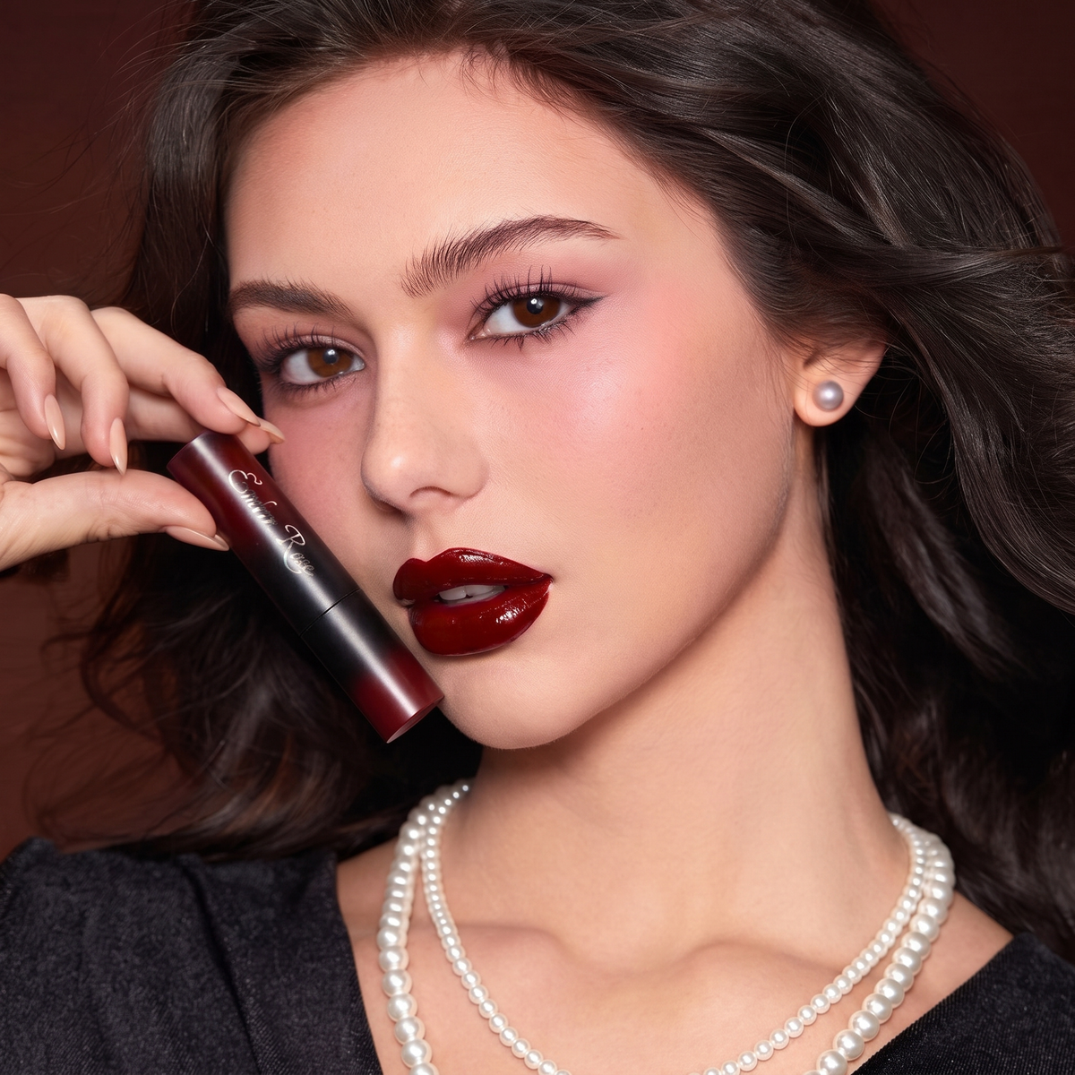A close-up portrait of a woman holding a dark red lip gloss tube near her face, wearing a deep glossy red lip, pearl necklace, and a single pearl stud earring against a warm brown background.